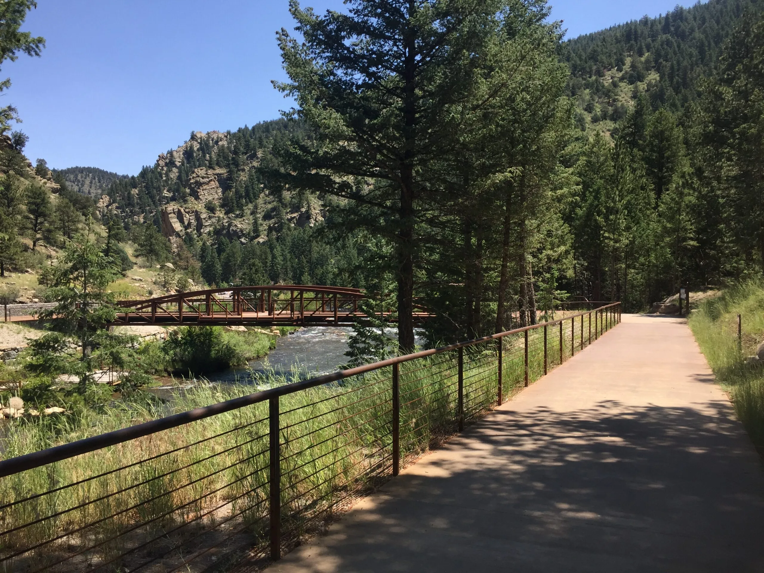 Golden’s creekside bike path crossing a rust-colored bridge toward pine-covered canyons.